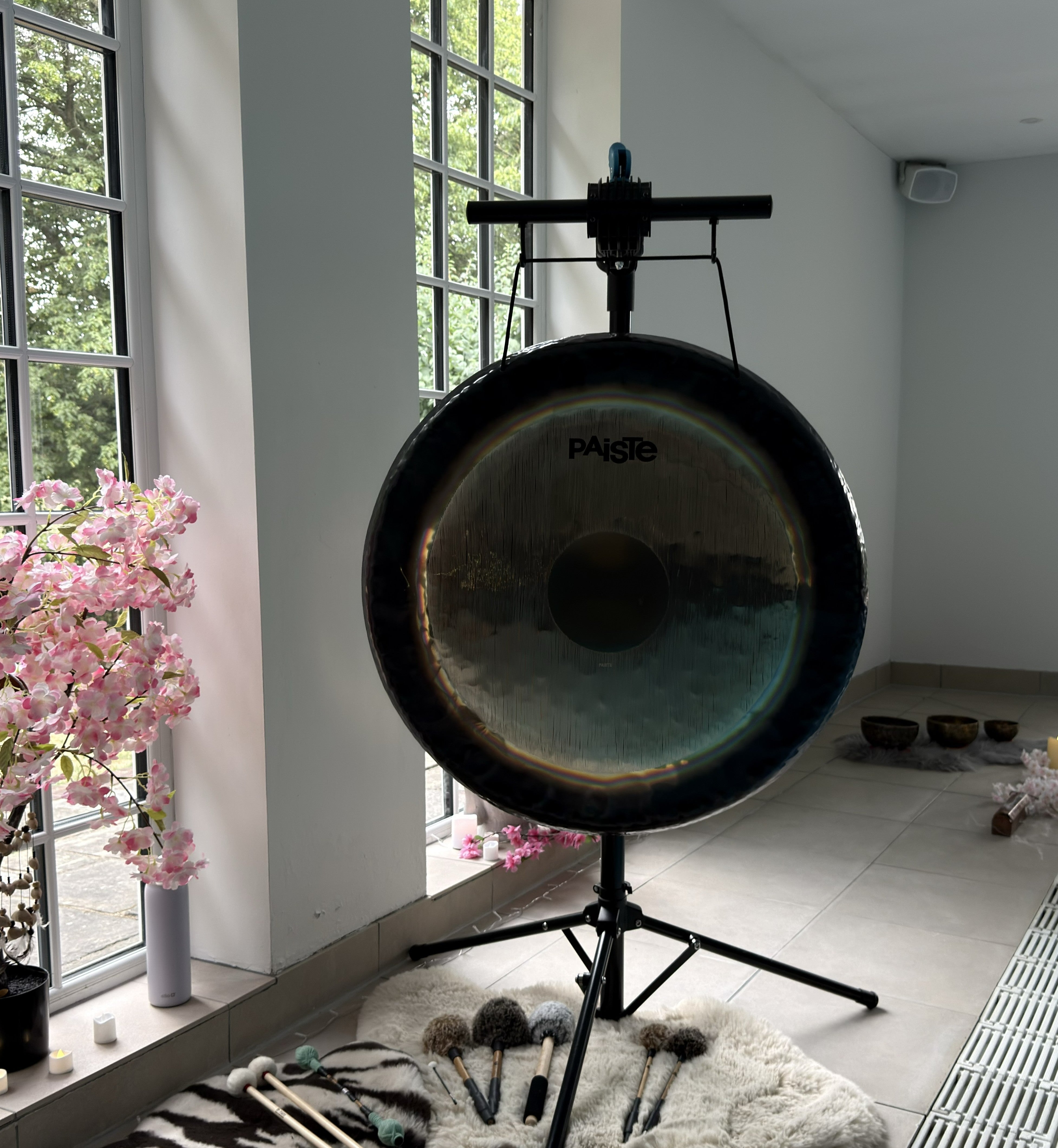 A large PAiSe gong on a black metal stand held by a Gong Bar inside a bright room with tall windows, pink cherry blossom decorations, and percussion mallets on a rug.